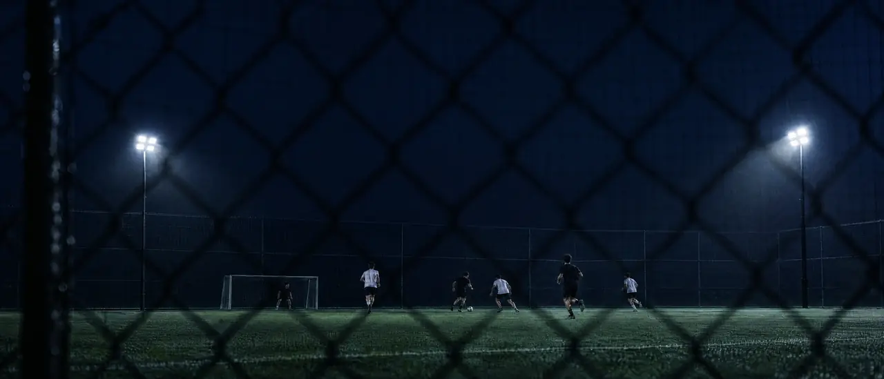A floodlit turf field at dusk seen through a black chain-link fence